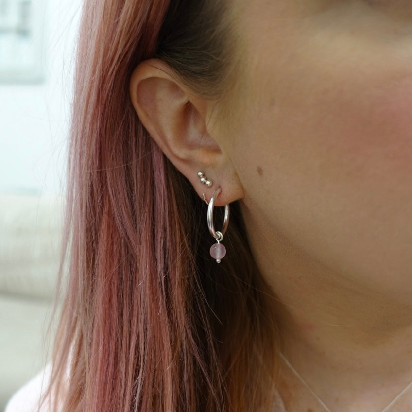 A close up shot of a pair of moon and dusk sterling silver hoop earrings with a delicate pink piece of rose quartz handing from them. Worn by a model