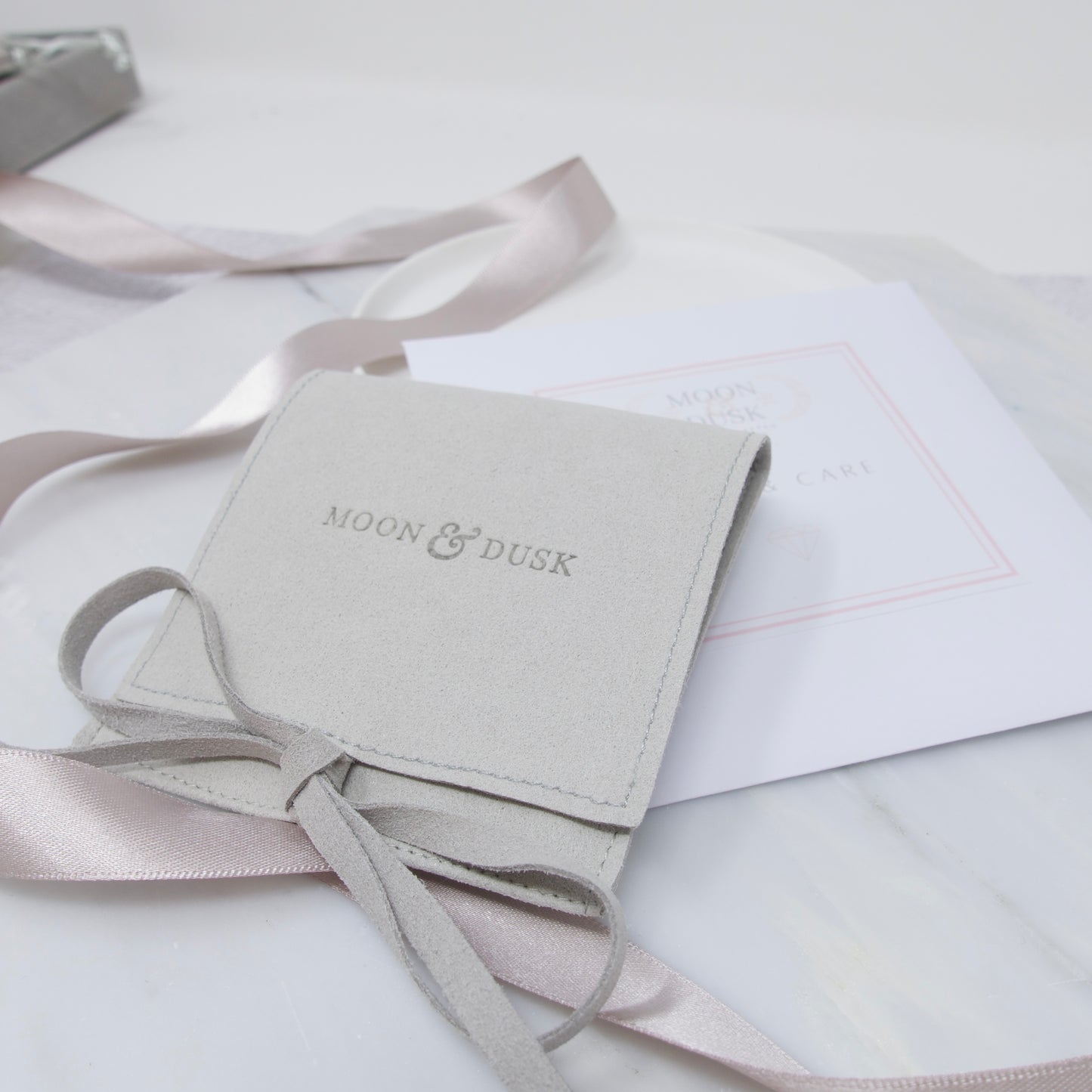 A soft grey Moon and Dusk jewellery pouch, tied with a bow, sitting on a care kit 