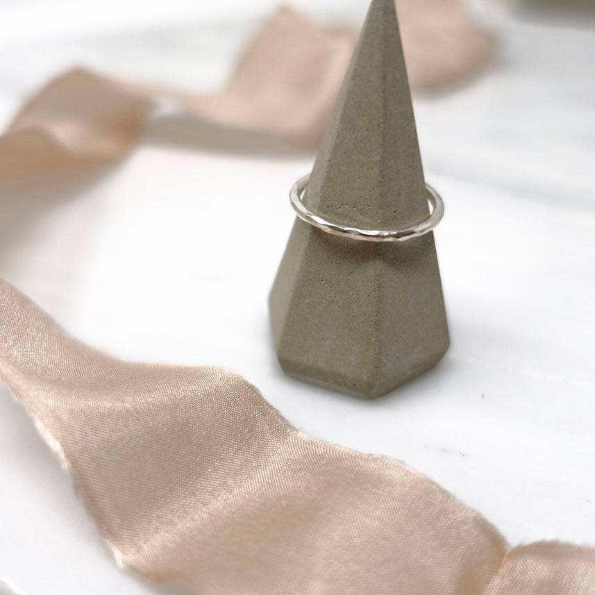 A ring cone and fraying piece of vintage pink silk ribbon have been placed on a marble background. Sitting on the ring cone is a sparkling and dainty sterling silver Moon and Dusk stacking ring